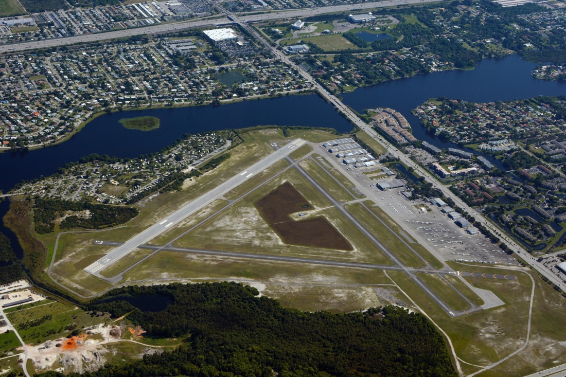 Aerial photograph taken above Palm Beach County Park Lantana Airport. Multiple numbered runways, airplane hangars, parked aircraft, and airport grounds are visible. Surrounding features include residential neighborhoods, wooded areas, roads, and part of Lake Osborne bordering the airport.