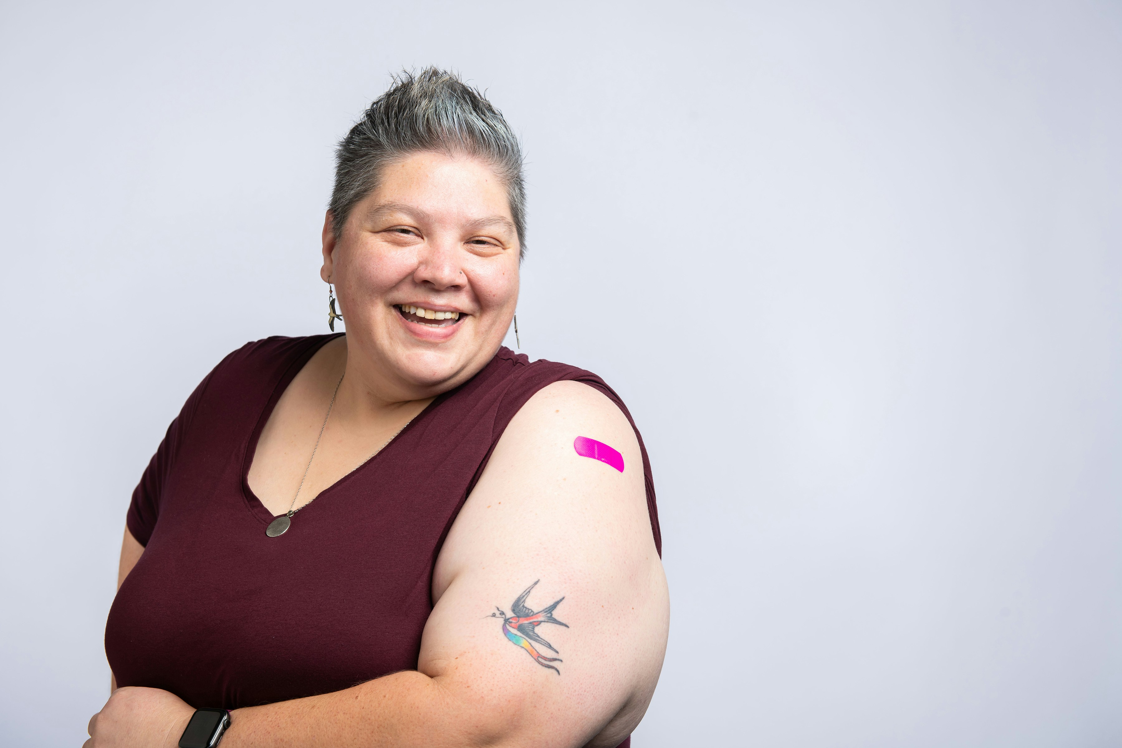 A smiling patient with short gray hair stands in a relaxed pose, wearing a maroon shirt. There is a pink adhesive bandage on their upper arm, just above a colorful bird tattoo.